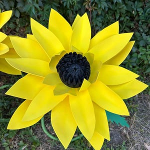May include: A large decorative flower sculpture featuring multiple layers of bright yellow, pointed petals arranged in a radial pattern around a textured, dark black circular center. The flower is supported by a thin green stem.