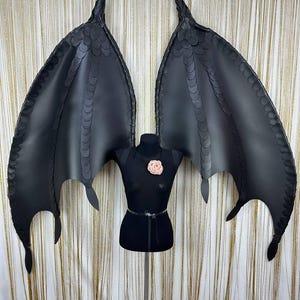 Puede incluir: Alas de drag&oacute;n negras de cuero sint&eacute;tico con un patr&oacute;n de escamas detallado. Las alas est&aacute;n unidas a un arn&eacute;s negro sobre un maniqu&iacute;. Una flor rosa adorna el pecho del maniqu&iacute;. Las alas est&aacute;n extendidas sobre un fondo de cortina dorada.