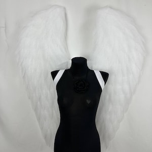 Puede incluir: Un maniquí negro con correas blancas y un collar de rosas negras, que muestra grandes alas de ángel blancas. Las alas están hechas de un material esponjoso y texturizado, y el maniquí está sobre un soporte metálico.
