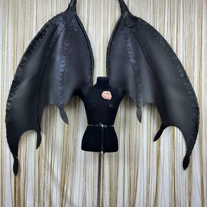 Puede incluir: Alas de drag&oacute;n negras de cuero sint&eacute;tico con un patr&oacute;n de escamas detallado. Las alas est&aacute;n unidas a un torso de maniqu&iacute; negro con un adorno floral rosa. Las alas est&aacute;n extendidas, mostrando su envergadura y dise&ntilde;o.