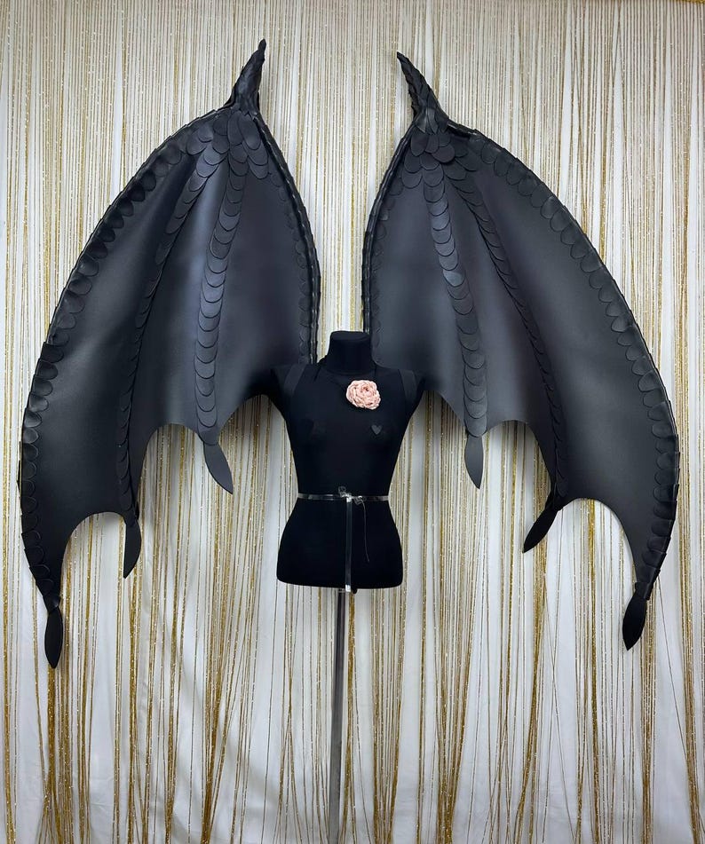 Puede incluir: Alas de drag&oacute;n negras de cuero sint&eacute;tico con un patr&oacute;n de escamas detallado. Las alas est&aacute;n unidas a un torso de maniqu&iacute; negro con un adorno floral rosa. Las alas est&aacute;n extendidas, mostrando su envergadura y dise&ntilde;o.