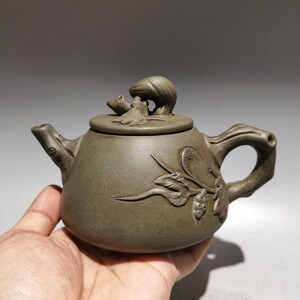 May include: A dark olive-green clay teapot featuring a sculpted frog perched on the lid. The body of the teapot is decorated with embossed leaf and vine motifs. It includes a curved spout, a sturdy handle, and a matching lid with a frog finial.