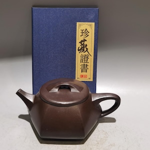 Op de afbeelding: Een donkerbruine zeshoekige theepot met bijpassend deksel en gebogen handvat, geplaatst voor een blauwe rechthoekige doos met een verticaal geel label met zwarte Chinese karakters en een klein rood zegel aan de onderkant.
