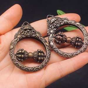 May include: Two circular metal pendants featuring an intricate relief of a mythical creature at the top and a central horizontal vajra symbol. The pendants have a dark, antiqued bronze finish with detailed scrollwork patterns along the outer ring.