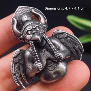 Pode incluir: Um pingente de metal prateado com uma figura alada e com chifres, com rosto de pássaro e detalhes texturizados. A figura segura dois longos fios de contas. O objeto mede 4,7 x 4,1 cm, conforme indicado pelo texto impresso na imagem.