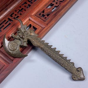 May include: A bronze-colored miniature axe with a dragon design. The axe head features a circular emblem, and the handle is shaped like a dragon's body. The axe is set against a dark wooden background.