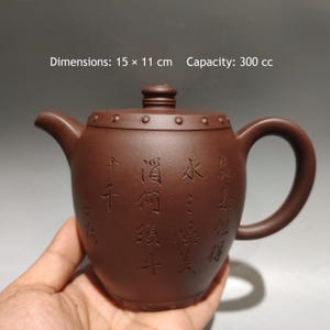 May include: A brown clay teapot with a curved handle and spout, featuring a lid with a small knob. The body is decorated with engraved Chinese characters and a row of small circular studs. Dimensions are 15 x 11 cm with a capacity of 300 cc.