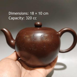 Puede incluir: Una tetera de arcilla marrón con cuerpo redondeado, pico curvo, asa y tapa con un pequeño pomo. La superficie presenta caracteres chinos grabados. El texto indica unas dimensiones de 18 x 10 cm y una capacidad de 320 cc para este recipiente.