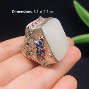 Peut inclure: Une bague argentée dotée d'une grande pierre blanche rectangulaire et lisse, sertie dans une monture en métal texturé. L'anneau présente des détails métalliques complexes avec des motifs d'oiseaux bleus et rouges. Dimensions : 3,1 x 2,2 cm.