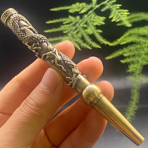 May include: A brass-colored, dragon-decorated pipe. The pipe features an ornate dragon design along its length and a decorative band at the top. The pipe tapers to a narrow end. The background is blurred with green foliage.