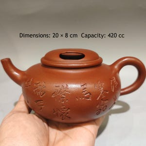 May include: A reddish-brown clay teapot featuring a rounded body, a curved spout, a handle, and a lid with a small opening. The exterior is decorated with carved Chinese characters. The text states dimensions of 20 x 8 cm and a capacity of 420 cc.