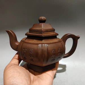 Puede incluir: Una tetera de arcilla marrón con cuerpo cuadrado, pico curvo y asa. El exterior presenta paneles verticales y caracteres chinos grabados. Tiene una tapa escalonada con un pequeño pomo y descansa sobre una base cuadrada.