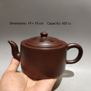 May include: A dark brown clay teapot with a cylindrical body, a curved spout, and a handle. The lid features a small knob and cut-out geometric patterns. Text on the image reads: Dimensions: 19 x 10 cm Capacity: 420 cc. The teapot has a matte finish.
