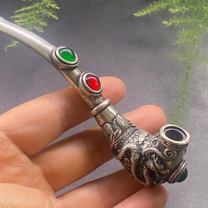 Pode incluir: Um cachimbo prateado com um motivo de dragão na tigela. O cachimbo apresenta uma gema verde e uma vermelha, e uma gema verde na base da tigela. A haste é longa e fina.