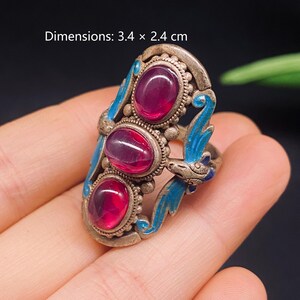 May include: A silver-toned ring featuring three oval-shaped, deep red cabochon stones arranged vertically. The metal band is decorated with blue enamel accents and intricate metalwork. The ring measures 3.4 x 2.4 cm as indicated by the text.