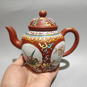 May include: A small, ornate teapot in reddish-brown with a lid. The teapot has hand-painted floral and abstract designs in white, yellow, and green. The handle and spout are curved, and the lid has a small knob.