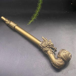 May include: A brass-colored pipe with a dragon head design. The pipe features intricate details, including scales and a detailed dragon head. The stem is cylindrical, and the bowl has a curved shape. The overall design is ornate and decorative.