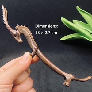 May include: A brown, elongated, animal-shaped figurine with a single curved horn, a ridged back, and a long tail. The object features a textured surface and measures 18 x 2.7 cm. The text Dimensions: 18 x 2.7 cm is printed on the image.