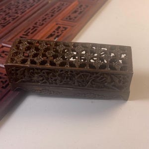 Può includere: Una scatola rettangolare in legno marrone scuro con intagli intricati e traforati sulla parte superiore e sui lati. La scatola presenta un motivo floreale decorativo e viene probabilmente utilizzata per la conservazione o l'esposizione. La scatola è su una superficie bianca.