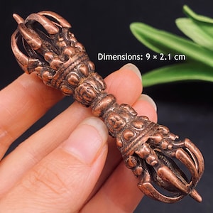 May include: A copper-coloured metal vajra ritual object featuring intricate embossed detailing and curved prongs at both ends. The object displays the text Dimensions: 9 x 2.1 cm, indicating its physical size in centimetres for reference.