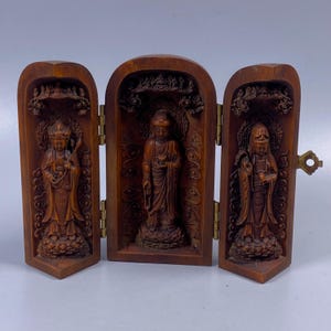 May include: A wooden triptych featuring three carved figures of Buddhist deities. The figures are in a standing pose, with detailed robes and serene expressions. The triptych is hinged and has a brass clasp. The wood has a warm, reddish-brown tone.