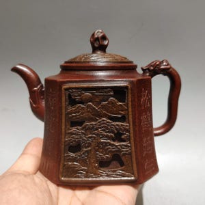 May include: A hexagonal brown clay teapot featuring an intricate openwork relief carving of a landscape with a tree and sun. The teapot has a curved spout, a dragon-shaped handle, and a lid with a sculpted finial. Chinese characters are engraved.