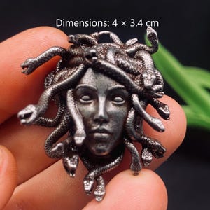 May include: A dark silver-toned metal pendant featuring a sculpted face surrounded by multiple snakes. The piece has a textured, antique finish and measures 4 x 3.4 cm. The design depicts a human-like face with snakes replacing hair.