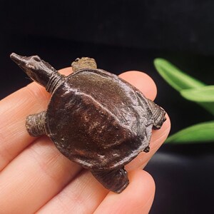 Op de afbeelding: Een kleine, donkerbruine metalen schildpadfiguur met een getextureerd, afgerond schild, een puntige kop en vier uitgestrekte ledematen. Het oppervlak van het object vertoont een verweerde, matte afwerking met zichtbare ribbels en contouren.