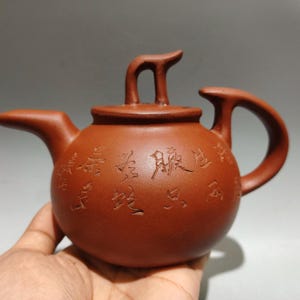 May include: A round, reddish-brown clay teapot featuring a curved spout, a handle with an upward extension, and a lid with a tall, arched finial. The body of the teapot is decorated with carved Chinese characters across the front surface.