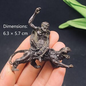 Peut inclure: Une figurine en métal de couleur bronze foncé représentant un personnage barbu chevauchant un grand félin. Le personnage a un bras levé vers le haut. L'objet mesure 6,3 cm sur 5,7 cm, comme indiqué par le texte imprimé sur l'image.