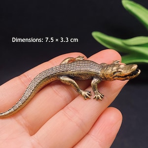 Könnte beinhalten: Eine kleine, metallene, bronzefarbene Alligatorfigur mit einer strukturierten, schuppigen Oberfläche und offenem Maul. Die Figur misst 7,5 mal 3,3 cm. Sie hat vier Beine mit sichtbaren Krallen und einen langen, geschwungenen Schwanz.