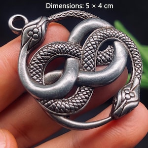 May include: A silver-toned metal pendant featuring two intertwined snakes forming a circular shape with textured scales and detailed heads. The pendant includes a small loop at the top for attachment and measures 5 x 4 cm.