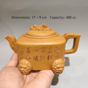 May include: A tan clay teapot with a square body, a lid featuring a sculpted creature, and four feet shaped like animal heads. The body displays engraved Chinese calligraphy. Dimensions are 17 x 9 cm with a capacity of 400 cc.