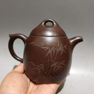 May include: A dark brown clay teapot featuring a rounded body with an engraved bamboo design on the front. The teapot has a curved handle, a short spout, and a lid with a circular handle opening at the top, showcasing a smooth matte finish.