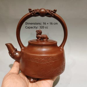 Könnte beinhalten: Eine braune Teekanne mit einem hohen, gebogenen Henkel, einem Ausguss und einem Deckel mit einer kleinen Tierfigur. Der Körper ist mit einer Landschaftsszene graviert. Der Text lautet: Dimensions: 16 x 16 cm, Capacity: 320 cc. Die Kanne ist matt.