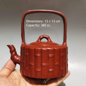 May include: A reddish-brown clay teapot with a bamboo-textured cylindrical body, a high arched handle, and a lid with a small loop handle. The teapot features printed text stating Dimensions: 13 x 13 cm and Capacity: 360 cc on the upper section.