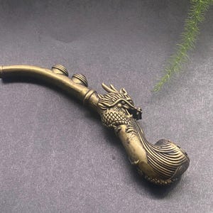 Può includere: Una pipa vintage in ottone con un design a testa di drago. La pipa ha un gambo curvo con tre piccoli dettagli circolari e una testa di drago dettagliata sulla testa. L'ottone ha un aspetto invecchiato e antico.