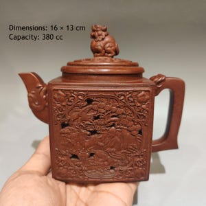 Op de afbeelding: Een bruine vierkante theepot met gedetailleerde reliëfsculpturen van figuren en bomen aan de zijkant, een deksel met een gebeeldhouwd wezen, een gebogen tuit en een handvat. De theepot meet 16 bij 13 cm en heeft een inhoud van 380 cc.
