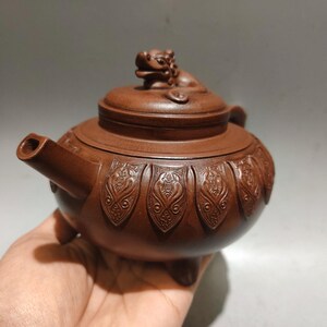 Può includere: Una teiera in argilla marrone con una figura scolpita di una creatura mitica sul coperchio. Il corpo della teiera è decorato con una serie di motivi in rilievo a forma di petalo attorno al centro ed è sostenuto da tre piccoli piedi.