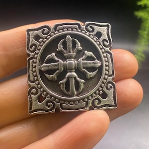 May include: A silver-coloured, square pendant with an elaborate design. The central feature is a detailed, symmetrical motif within a circular frame, surrounded by a decorative border. The pendant is held in a person's hand.