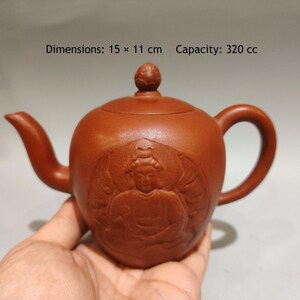 Könnte beinhalten: Eine rotbraune Tonteekanne mit einem Relief einer sitzenden Figur auf der Vorderseite. Die Teekanne hat einen Deckel mit einem kleinen Knauf, einen geschwungenen Ausguss und einen Henkel. Der Text lautet: Dimensions: 15 x 11 cm Capacity: 320 cc.
