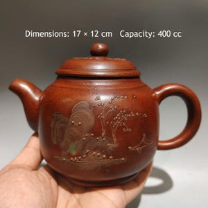 May include: A brown clay teapot with a lid, featuring an embossed landscape design on the body. The teapot measures 17 x 12 cm and has a capacity of 400 cc. The surface has a matte texture with a curved handle and a short spout.