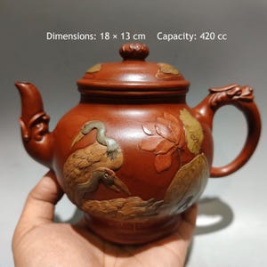 May include: A reddish-brown clay teapot featuring relief carvings of a crane and lotus flowers. The teapot has a lid with a knob, a curved spout, and a handle shaped like a dragon. Text on the image states: Dimensions: 18 x 13 cm Capacity: 420 cc.