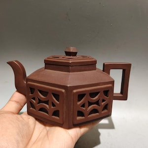 Puede incluir: Una tetera hexagonal de arcilla marrón con un intrincado patrón de celosía calada en sus lados. La tetera tiene una tapa plana con un pequeño pomo, un pico curvo y un asa rectangular, todo elaborado con un material cerámico mate y terroso.