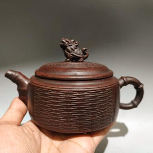 Peut inclure: Une théière en argile brun foncé avec un corps texturé à lignes horizontales, une anse en forme de bambou et un bec verseur. Le couvercle est surmonté d'une petite figurine de créature mythique sculptée et présente un motif circulaire.