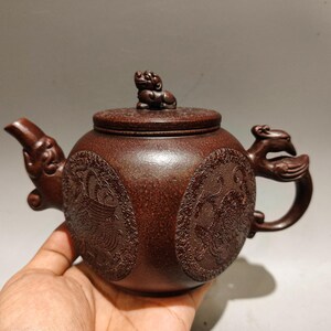 May include: A dark brown textured clay teapot featuring a round body with circular relief carvings of mythical creatures. The lid is topped with a small sculpted lion figure, while the spout and handle are shaped like stylized animal heads.