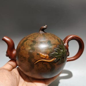 May include: A round brown teapot with a mottled green surface texture resembling a watermelon. The teapot features a curved spout, a looped handle, and a small stem-shaped lid knob. A sculpted green leaf and a small yellow vine are attached.
