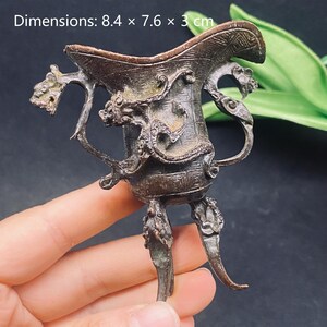 Könnte beinhalten: Ein dunkel bronzefarbenes Metallgefäß mit drei Beinen und einem ausgestellten Rand, das auf der Außenseite mit komplexen Drachenreliefs verziert ist. Das Objekt zeigt ein gitterartiges Muster und misst laut Text 8,4 x 7,6 x 3 cm.