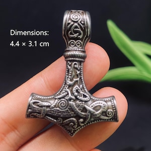 May include: A silver-toned metal pendant shaped like a hammer, featuring intricate embossed knotwork and spiral patterns. The pendant measures 4.4 by 3.1 cm as indicated by the text. It has a textured, oxidized finish with a bail at the top.