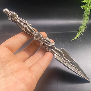 May include: A silver-colored dagger with an ornate handle and a dragon head design. The blade is pointed and features etched details. The dagger is held in a hand against a dark background.